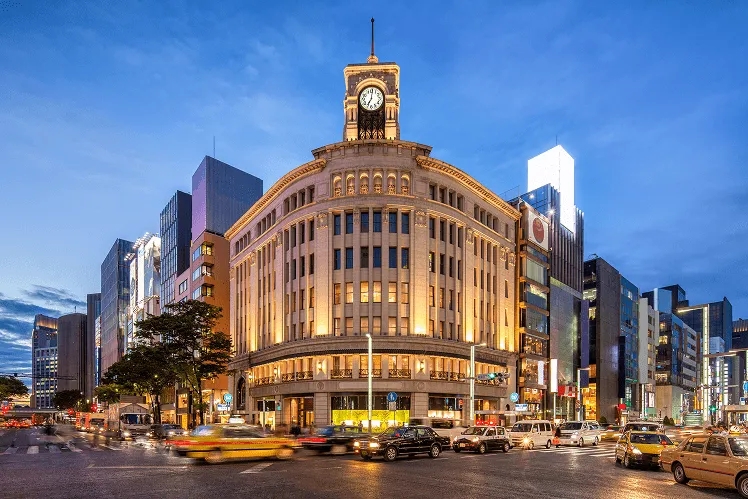 A vibrant evening with taxis maneuvering through the bustling streets in Ginza, Tokyo, around Wako department store featuring its iconic clock tower against the dark blue sky