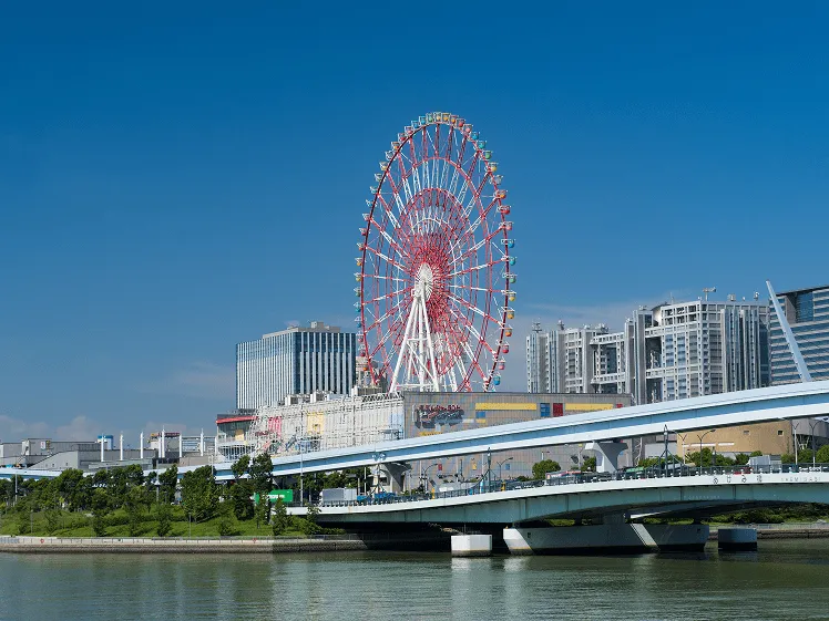 A bright day near Tokyo Bay with spectacular views of the city skyline and river, featuring the iconic Palette Town Ferris Wheel standing vibrant and colorful against the clear blue sky