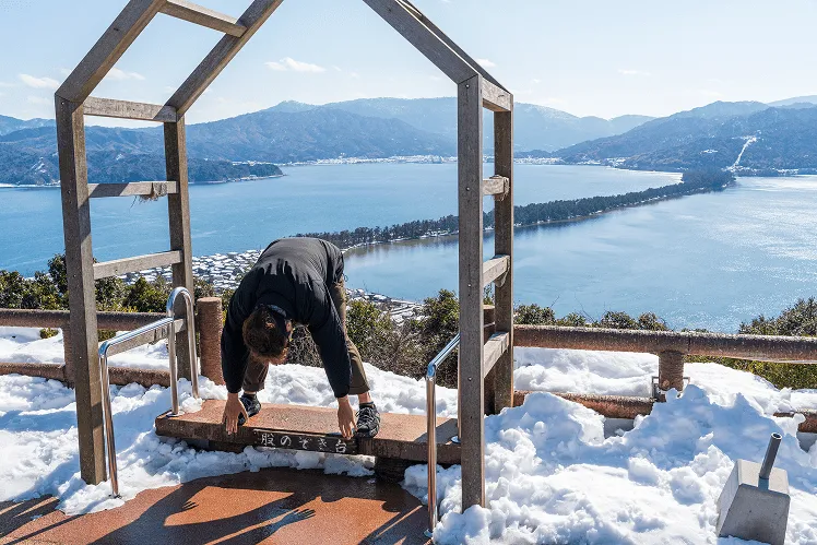 A man in winter stands at the Matanozoki viewpoint, bending over and looking through his legs toward the pine-covered sandbar stretching across Miyazu Bay