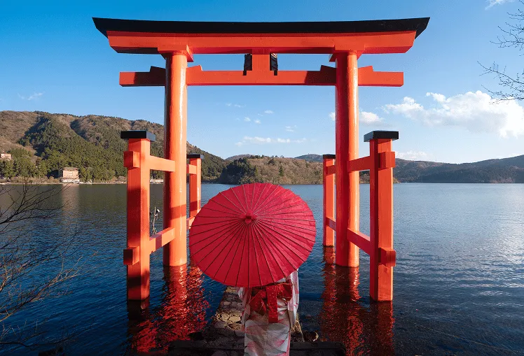 Standing in front of Miyajima: The Red Torii Gate on the Water, a woman dressed in traditional Japanese attire, wearing a kimono, gracefully holding a vibrant red umbrella