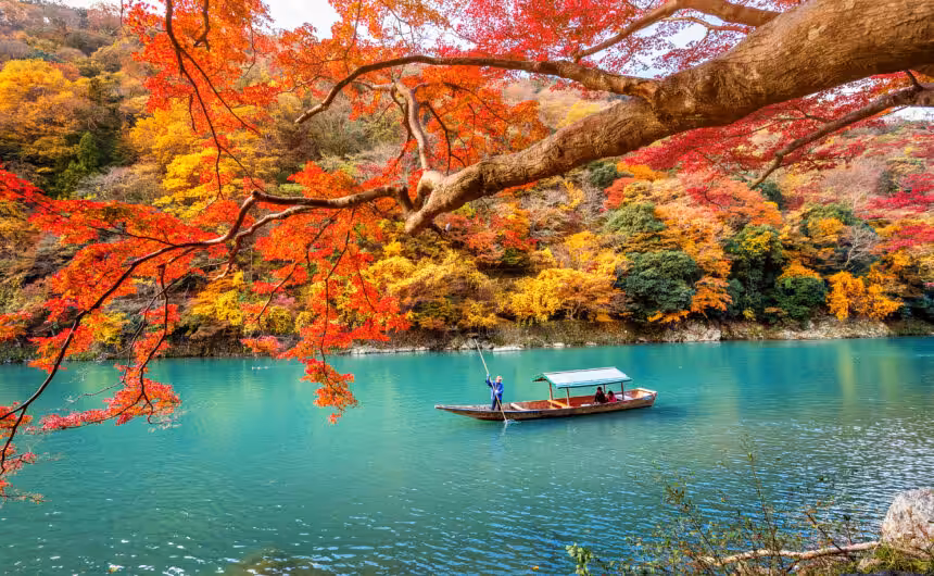 A boatman punts a wooden boat along the crystal-clear emerald river of Arashiyama, surrounded by vibrant autumn foliage in Kyoto, Japan